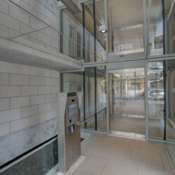 Modern building entrance with large glass doors, gray brick walls, and a stainless steel security kiosk near the door.