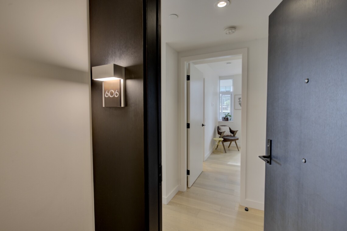 Open doorway into a bright living area; hallway with a dark wall and apartment number 606 plaque visible next to a lighted wall sconce by the door.