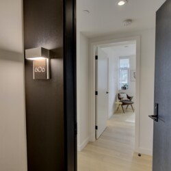 Open doorway into a bright living area; hallway with a dark wall and apartment number 606 plaque visible next to a lighted wall sconce by the door.