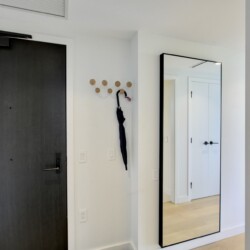 Interior hallway with a dark front door on the left, wooden coat hooks and an umbrella, and a tall framed mirror reflecting a white door.