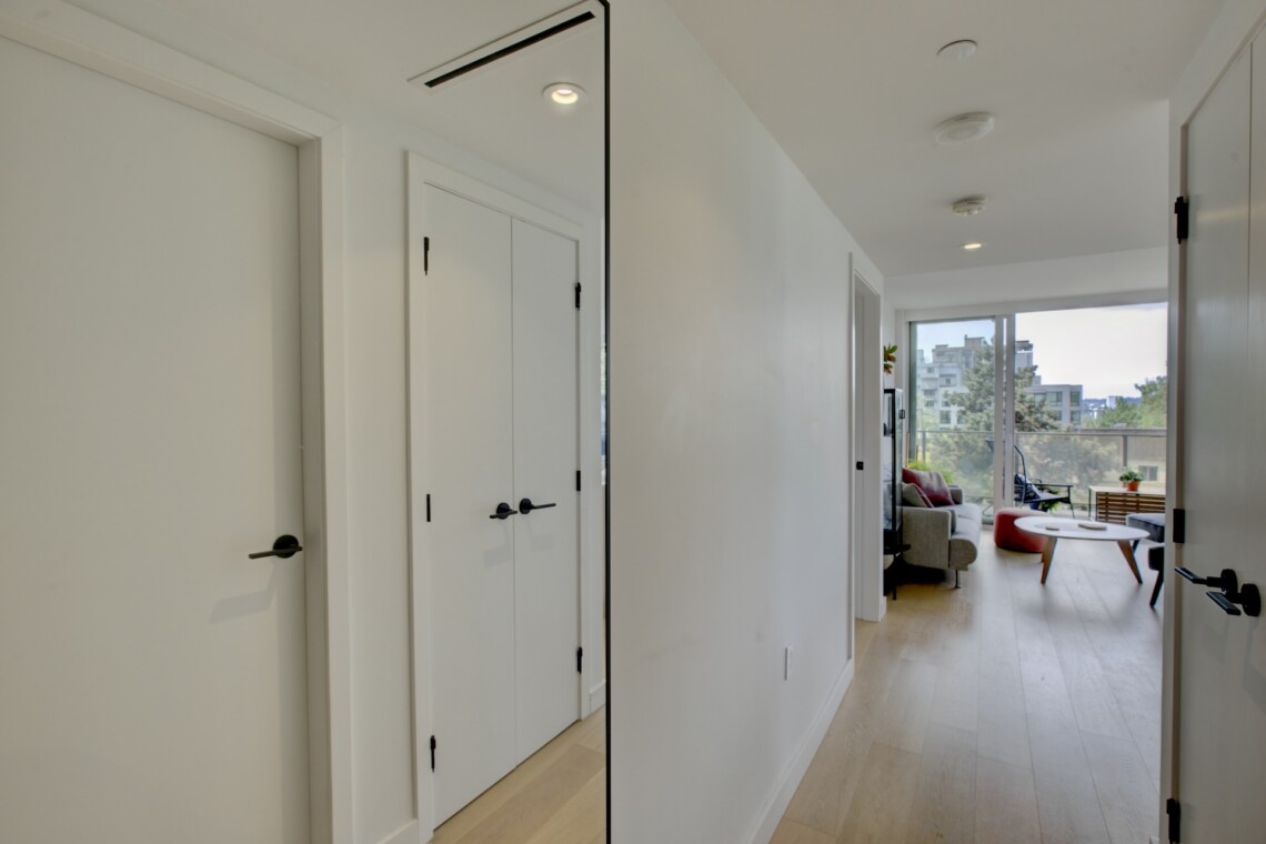 Bright white hallway with closed doors on the left and a sunny living room visible at the far end through a balcony doorway, wooden floor.
