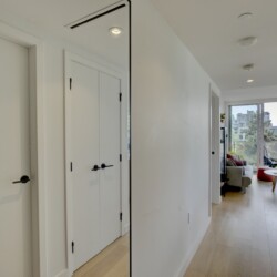 Bright white hallway with closed doors on the left and a sunny living room visible at the far end through a balcony doorway, wooden floor.