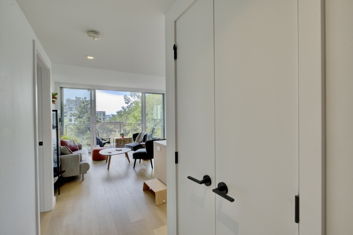 View from a hallway into a bright, modern living room with large windows and a balcony overlooking trees and buildings.