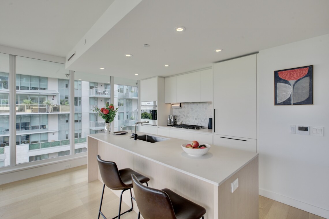 Bright, modern kitchen with white cabinets, an island, and large windows overlooking an urban cityscape with balconies and glass buildings.