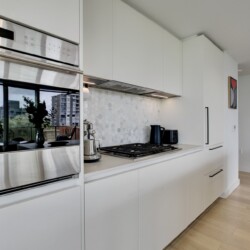 Modern white kitchen with stainless steel wall ovens, gas cooktop, and a hexagonal tiled backsplash between white cabinets.