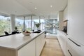 Contemporary white kitchen with an island, fruit bowl, vase of flowers, and large windows overlooking a cityscape and trees.