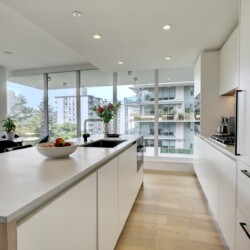 Contemporary white kitchen with an island, fruit bowl, vase of flowers, and large windows overlooking a cityscape and trees.