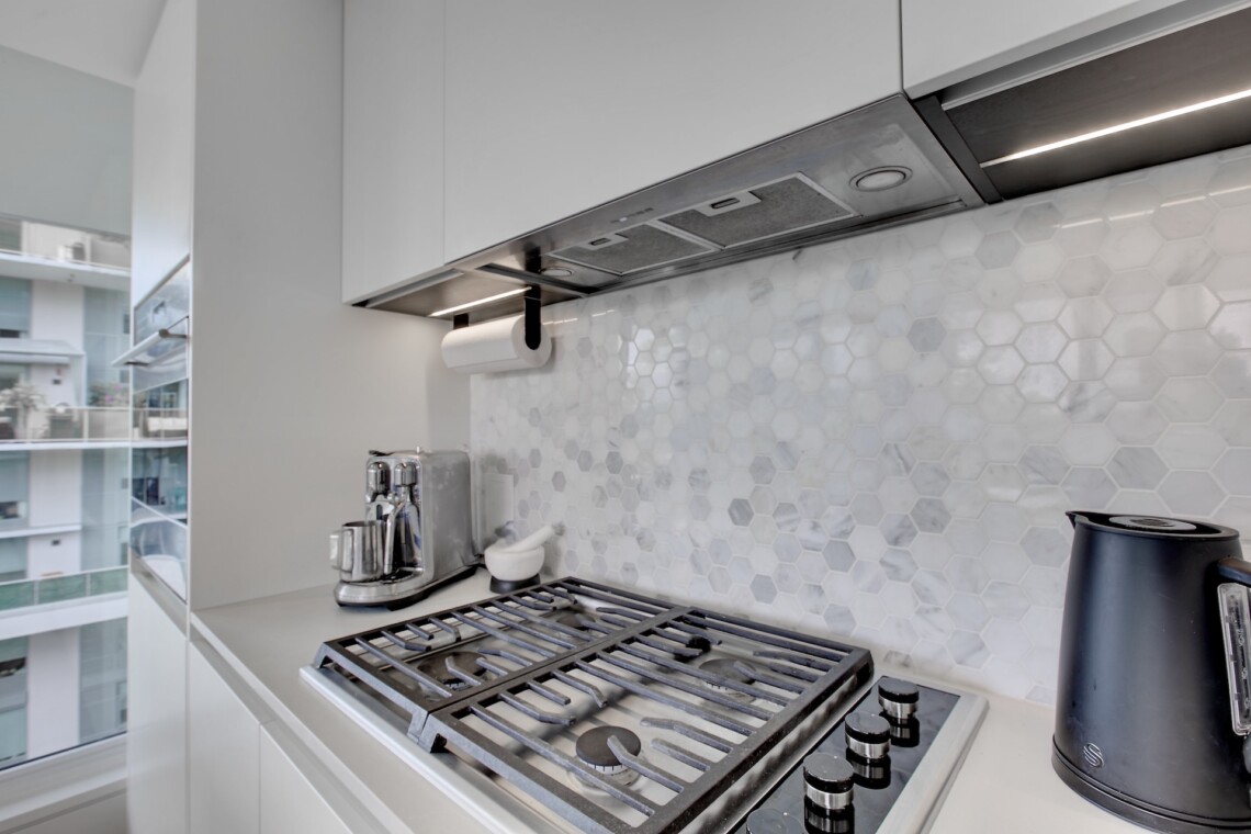 Modern kitchen with a gas range, stainless vent hood, and hexagonal marble backsplash; windowed city view in the background.