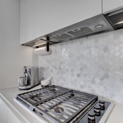 Modern kitchen with a gas range, stainless vent hood, and hexagonal marble backsplash; windowed city view in the background.