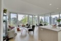 Bright, open-plan living area with panoramic glass walls; modern seating, round coffee tables, and a kitchen island overlooking a balcony and trees outside.