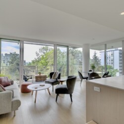 Bright, open-plan living area with panoramic glass walls; modern seating, round coffee tables, and a kitchen island overlooking a balcony and trees outside.