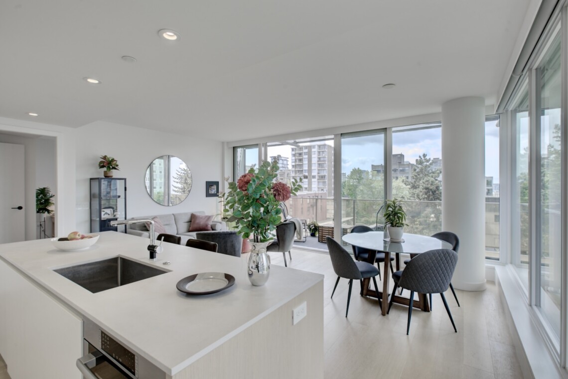 Open-plan living area with a white kitchen island, sink, and built-in oven in foreground; living room with gray sofa, round mirror, plants, and large glass windows overlooking cityscape.