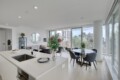 Open-plan living area with a white kitchen island, sink, and built-in oven in foreground; living room with gray sofa, round mirror, plants, and large glass windows overlooking cityscape.