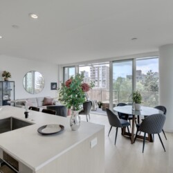 Open-plan living area with a white kitchen island, sink, and built-in oven in foreground; living room with gray sofa, round mirror, plants, and large glass windows overlooking cityscape.
