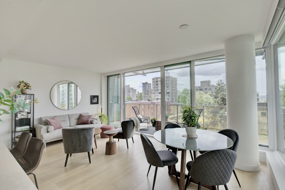 Bright, modern living area with a grey sofa, round wall mirror, plants, and a glass dining table near floor-to-ceiling windows showing a cityscape.