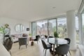 Bright, modern living area with a grey sofa, round wall mirror, plants, and a glass dining table near floor-to-ceiling windows showing a cityscape.