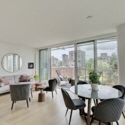 Bright, modern living area with a grey sofa, round wall mirror, plants, and a glass dining table near floor-to-ceiling windows showing a cityscape.