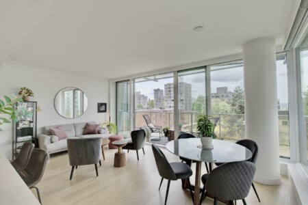 Bright, modern living area with a grey sofa, round wall mirror, plants, and a glass dining table near floor-to-ceiling windows showing a cityscape.