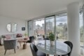 Bright modern living area with a gray sofa, pink cushions, round mirror, and glass dining table by large windows.