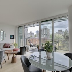 Bright modern living area with a gray sofa, pink cushions, round mirror, and glass dining table by large windows.