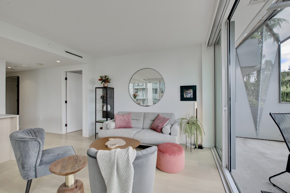 Bright modern living room with a gray sofa and pink cushions, circular wall mirror, and glass sliding doors to a balcony.
