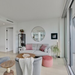 Bright modern living room with a gray sofa and pink cushions, circular wall mirror, and glass sliding doors to a balcony.