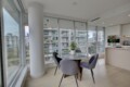 Bright, modern dining area with a round glass table and four gray upholstered chairs, overlooking a cityscape through large floor-to-ceiling windows; white cabinetry and a plant in the scene.
