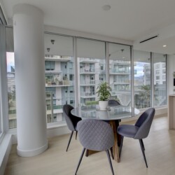 Bright, modern dining area with a round glass table and four gray upholstered chairs, overlooking a cityscape through large floor-to-ceiling windows; white cabinetry and a plant in the scene.