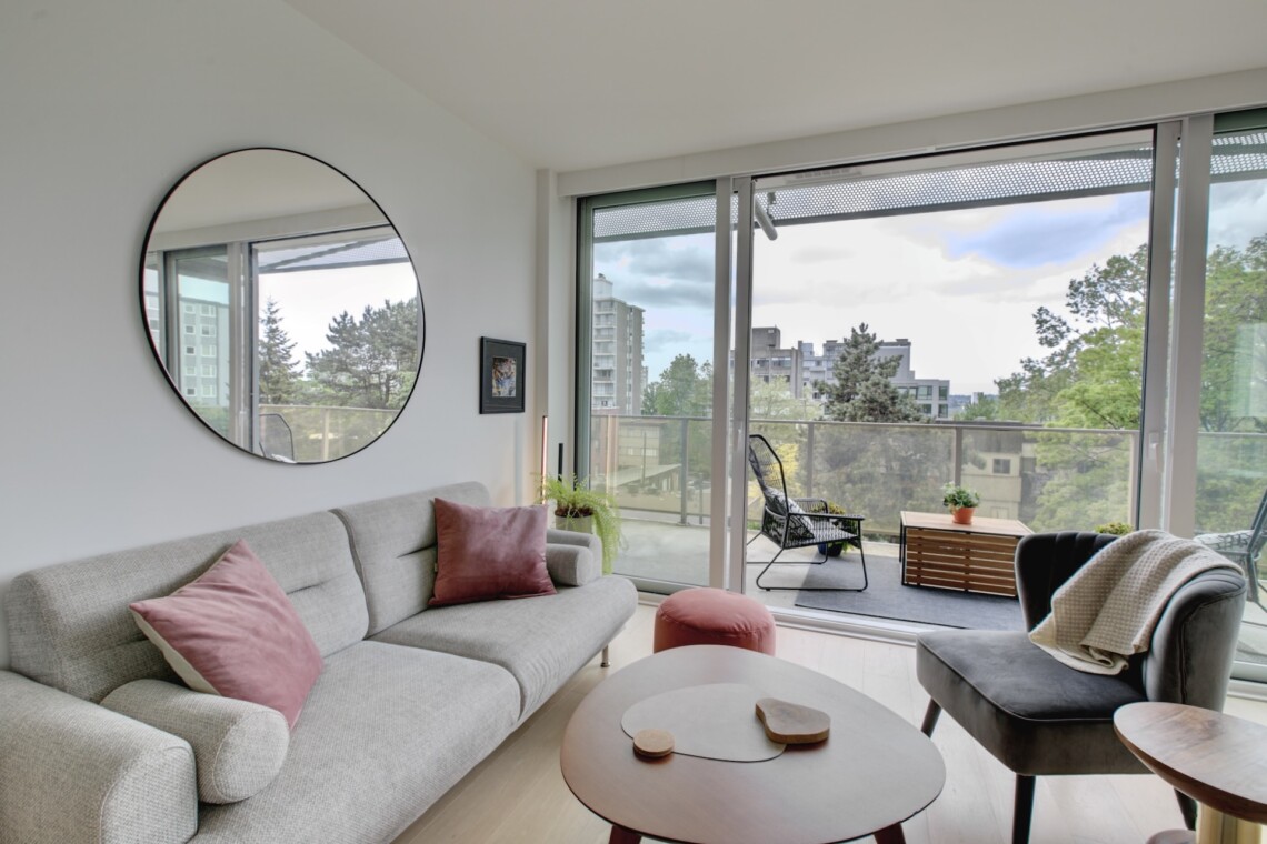 Bright living room with a round wall mirror, pale sofa with pink cushions, and a glass balcony door revealing an outdoor seating area and city view.