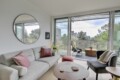 Bright living room with a round wall mirror, pale sofa with pink cushions, and a glass balcony door revealing an outdoor seating area and city view.