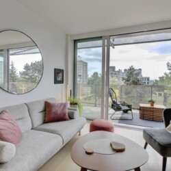 Bright living room with a round wall mirror, pale sofa with pink cushions, and a glass balcony door revealing an outdoor seating area and city view.