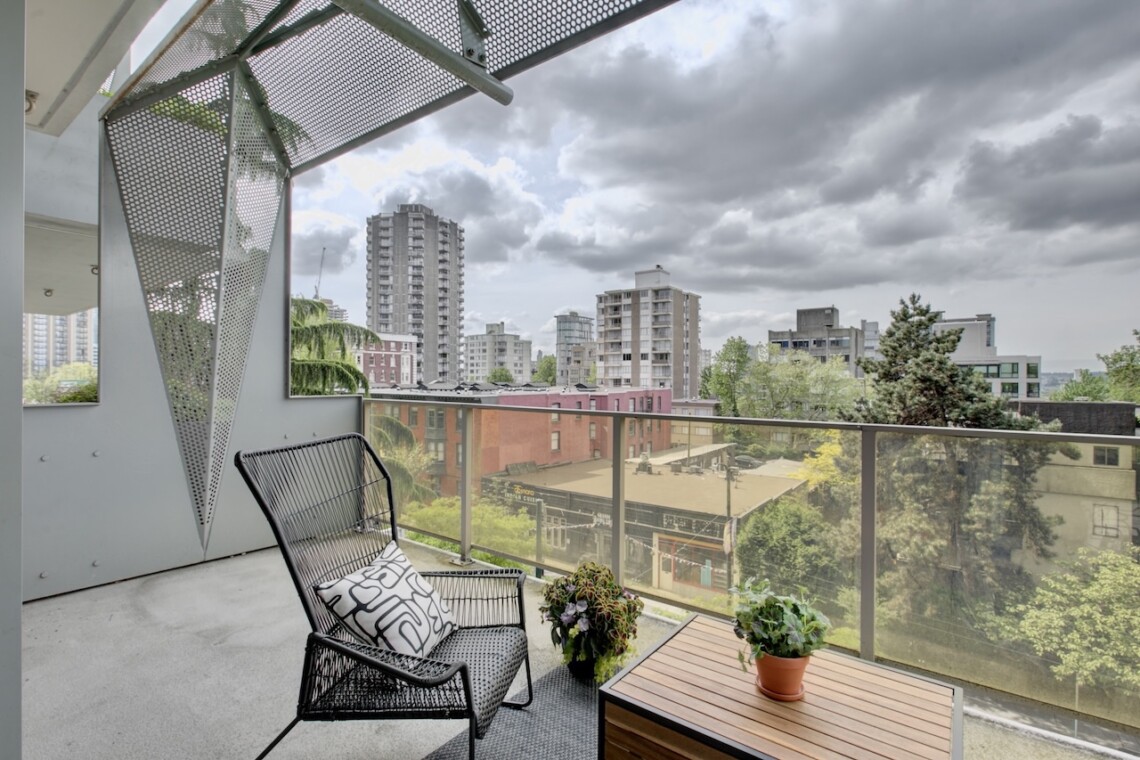 Balcony with a black metal chair, wooden table, and potted plants overlooking a city skyline behind a glass railing under a cloudy sky.",