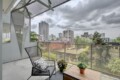 Balcony with a black metal chair, wooden table, and potted plants overlooking a city skyline behind a glass railing under a cloudy sky.",