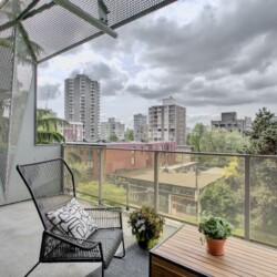 Balcony with a black metal chair, wooden table, and potted plants overlooking a city skyline behind a glass railing under a cloudy sky.",