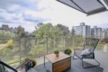 Balcony seating area with two black wire chairs, a wooden table, and potted plants, overlooking a tree-lined urbanscape.