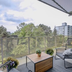 Balcony seating area with two black wire chairs, a wooden table, and potted plants, overlooking a tree-lined urbanscape.