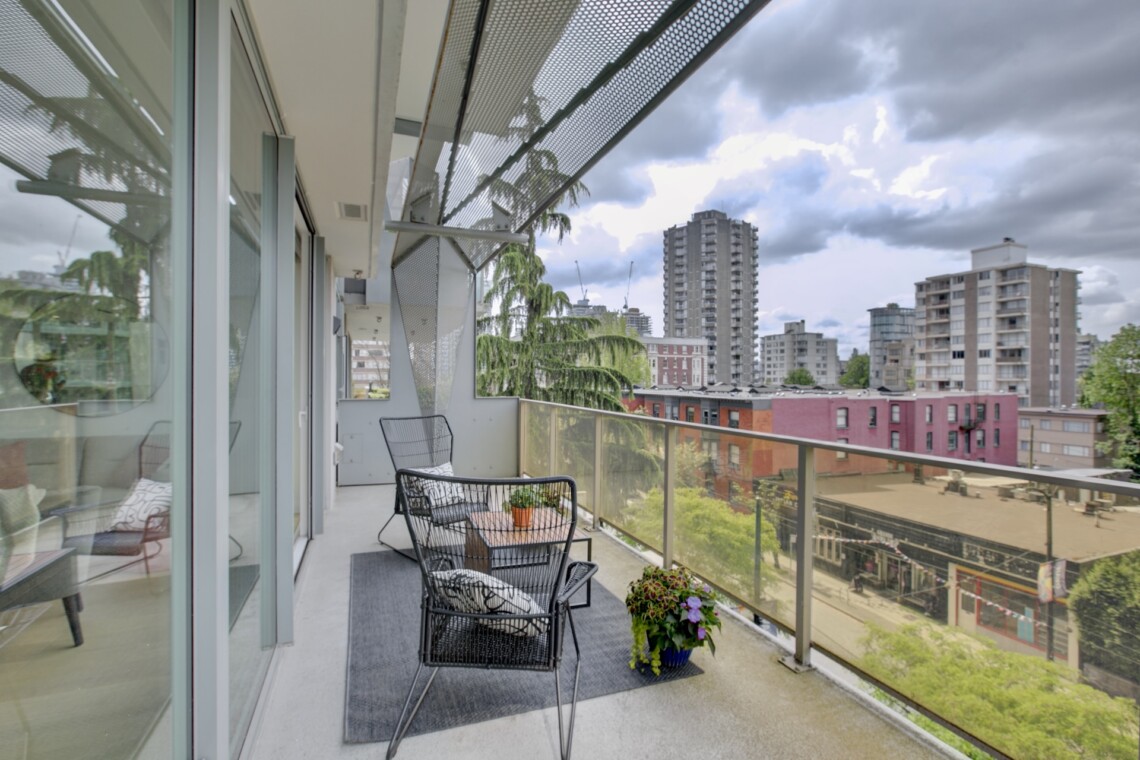 Balcony seating area with black wire chairs and a small table, potted plant, glass railing, overlooking a cityscape of mid-rise buildings under a cloudy sky.