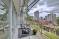 Balcony seating area with black wire chairs and a small table, potted plant, glass railing, overlooking a cityscape of mid-rise buildings under a cloudy sky.