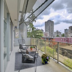 Balcony seating area with black wire chairs and a small table, potted plant, glass railing, overlooking a cityscape of mid-rise buildings under a cloudy sky.