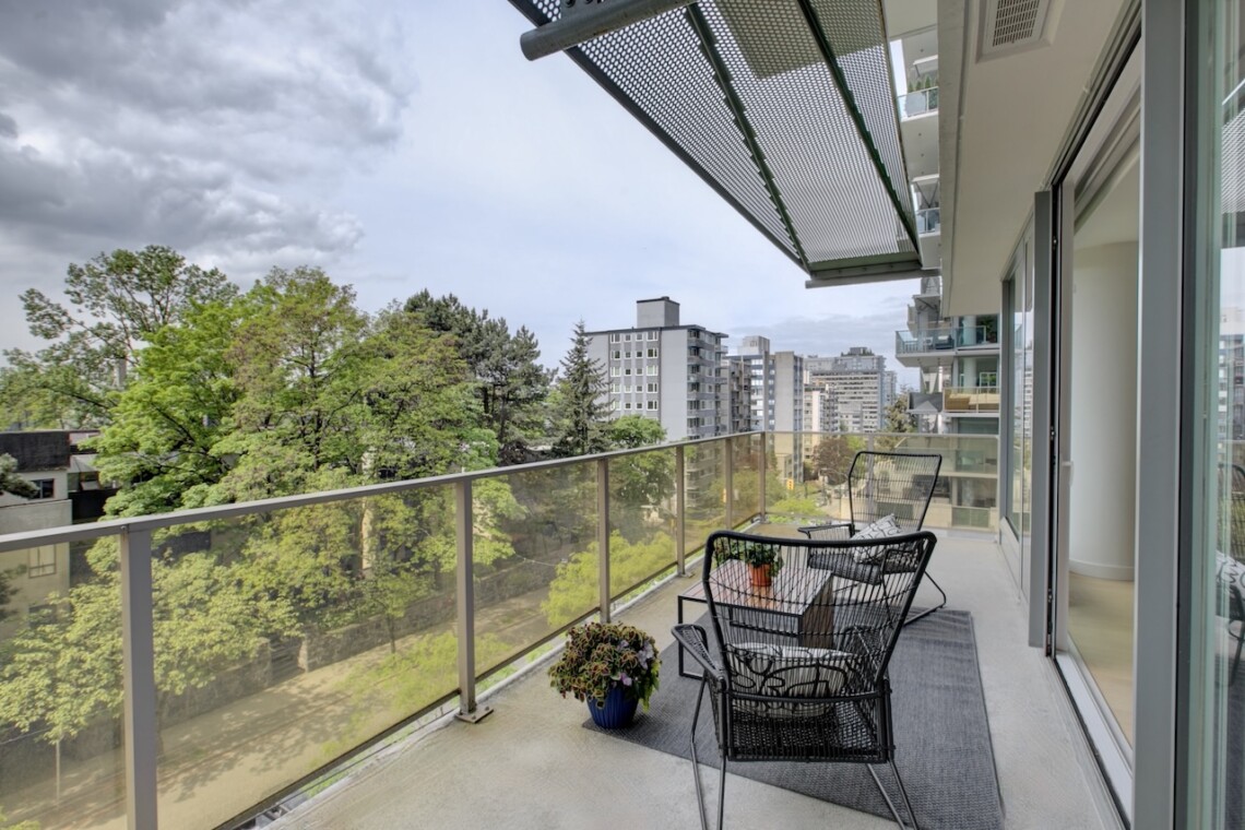 Balcony seating with two black wire chairs and a small table, glass railing, overlooking trees and city buildings in the distance.