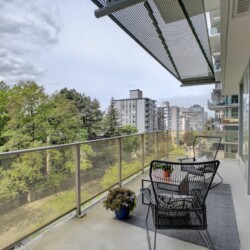 Balcony seating with two black wire chairs and a small table, glass railing, overlooking trees and city buildings in the distance.