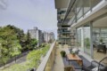 Balcony of a modern apartment overlooking a tree-lined street with other high-rise buildings in the distance, outdoor seating and potted plants visible