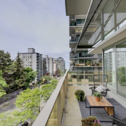 Balcony of a modern apartment overlooking a tree-lined street with other high-rise buildings in the distance, outdoor seating and potted plants visible