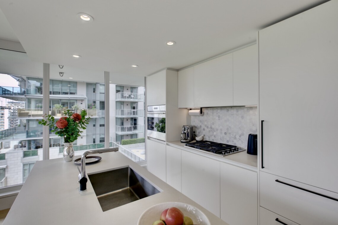 Bright modern white kitchen with an island, sink, and gas stove, overlooking a city view through large windows with a vase of flowers and a bowl of fruit in the foreground.