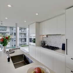 Bright modern white kitchen with an island, sink, and gas stove, overlooking a city view through large windows with a vase of flowers and a bowl of fruit in the foreground.