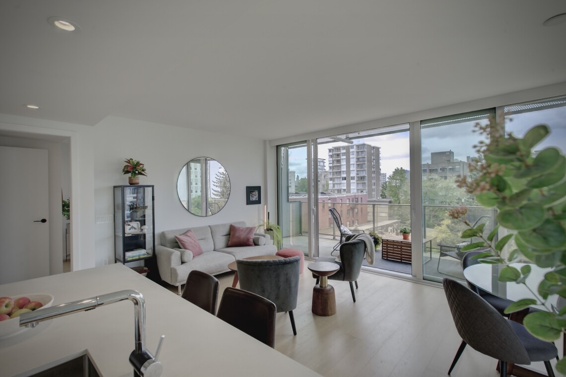 Bright living room with white walls, gray sofa, round mirror, and glass doors to a balcony overlooking the city.