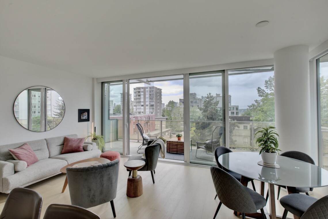 Bright modern living room with a gray sectional, pink cushions, and a round mirror, fronted by tall glass doors opening to a balcony with a city view.] ,