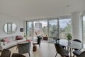 Bright modern living room with a gray sectional, pink cushions, and a round mirror, fronted by tall glass doors opening to a balcony with a city view.] ,