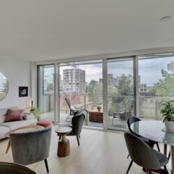 Bright modern living room with a gray sectional, pink cushions, and a round mirror, fronted by tall glass doors opening to a balcony with a city view.] ,