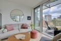 Bright living room with a gray sectional, pink cushions, round wall mirror, and glass balcony doors overlooking city buildings.